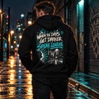Back of the black MUD After Dark Zip Hoodie showing the When The Days Get Darker, Speak Louder graphic, worn on a wet city street at night